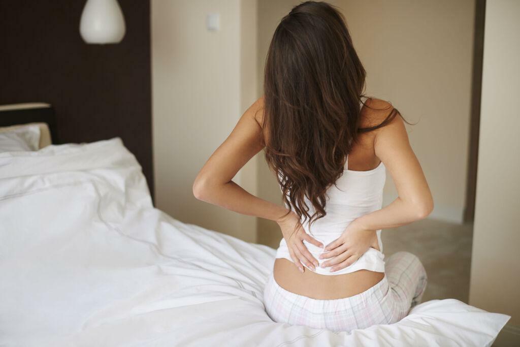 A person sitting at the edge of a bed, holding their back in pain.
